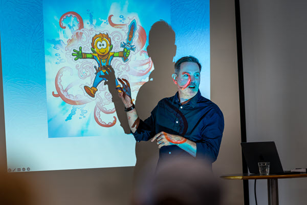 En oplægsholder står foran en projekter og fortæller om en tegnet figur (nomerne).